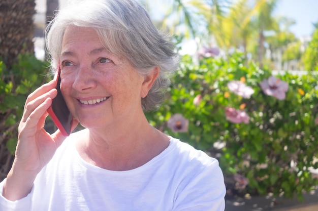 Attraktive ältere Frau lächelt im Gespräch mit dem Handy im Freien in einem grünen öffentlichen Park