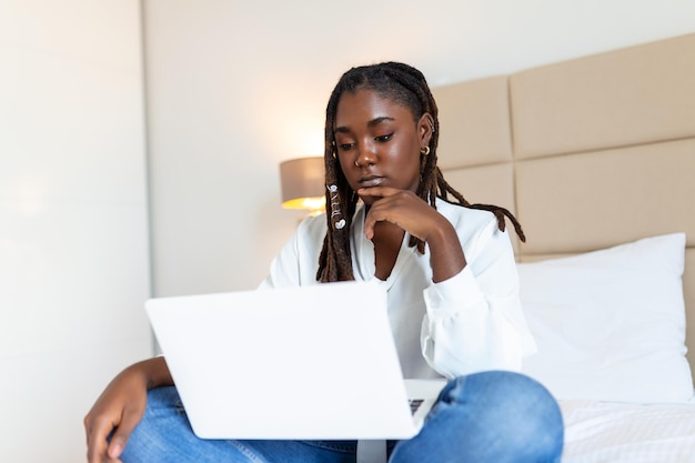 Attraktive afrikanische Frau in weißem Hemd mit Laptop und lächelnd am Morgen Video-Chat und freiberufliche Arbeit von zu Hause aus Konzept einer schönen jungen Frau mit Laptop im Bett