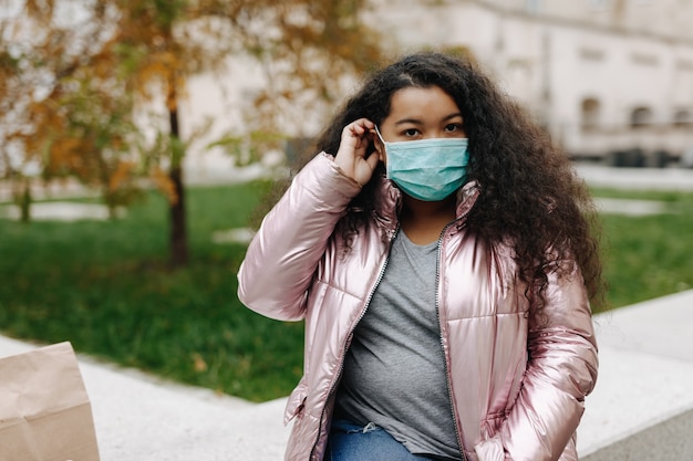 Attraktive afrikanische Frauen in der medizinischen Gesichtsmaske, die auf Straße aufwirft. Schutz vor Coronavirus. Pandemiezeit und Quarantäne.