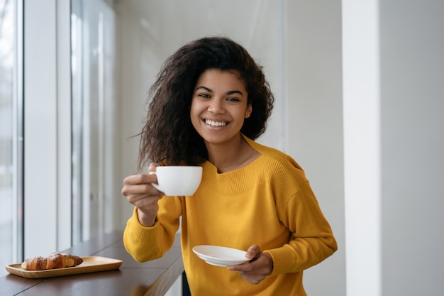 Attraktive afroamerikanische Frau, die Tasse Kaffee hält, Kamera betrachtet und lächelt