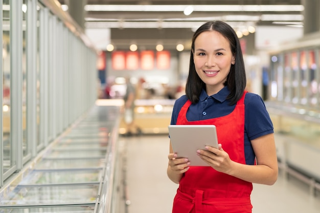 Attraktive Asiatin in Uniform mit roter Schürze, die im Supermarkt mit digitalem Tablet arbeitet