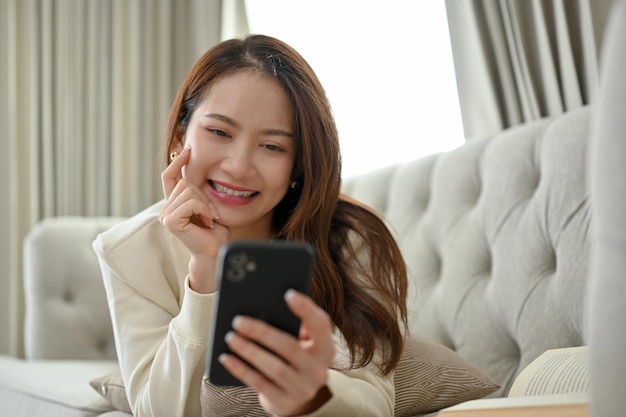 Attraktive asiatische Frau, die auf dem Sofa liegt und mit ihren Freunden auf ihrem Smartphone chattet