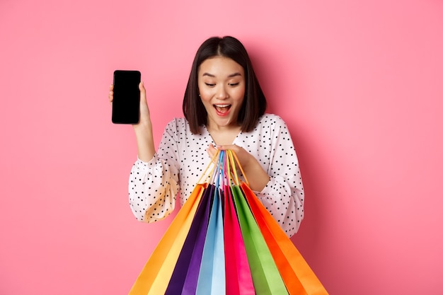 Attraktive asiatische Frau, die Smartphone-App und Einkaufstaschen zeigt, online über Anwendung kaufend, über rosa stehend