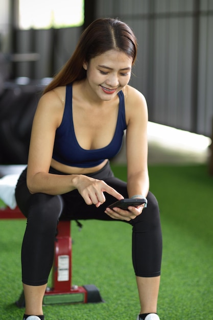 Attraktive asiatische Frau sitzt in einem Fitnessstudio und scrollt nach dem Training mit ihrem Smartphone in den sozialen Medien