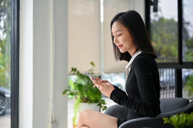 Attraktive asiatische Geschäftsfrau im schwarzen Kleid, die ihr Telefon benutzt, während sie im Café sitzt