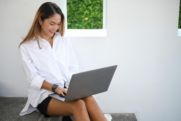 Attraktive asiatische junge Frau in einem lässigen Outfit, das auf der Treppe sitzt und ihren Laptop-Computer benutzt