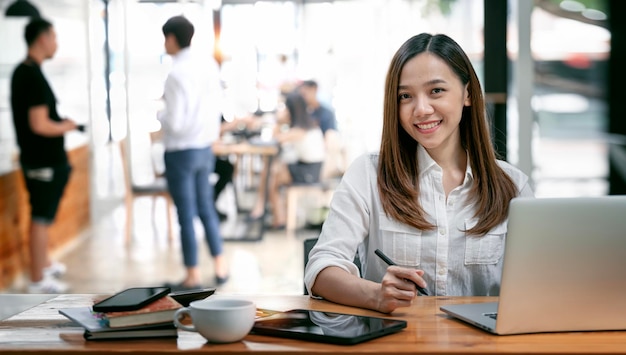 Attraktive asiatische junge selbstbewusste Geschäftsfrau sitzt am Bürotisch mit einer Gruppe von Kollegen im Hintergrund, die an einem Laptop arbeiten