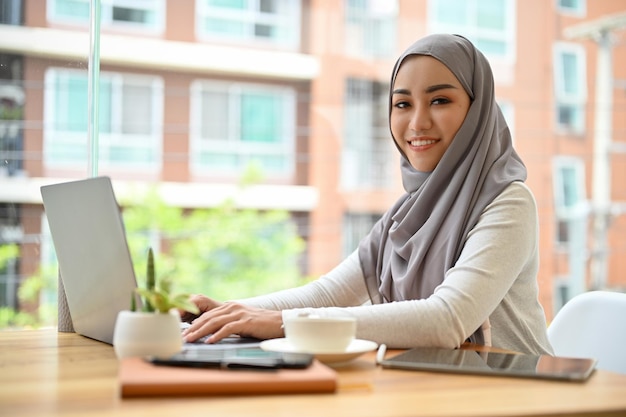 Attraktive asiatische muslimische Frau mit Hijab mit einem tragbaren Laptop-Computer im Büro