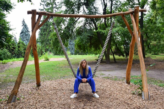 Attraktive blonde Frau, die auf einer hängenden Seilschaukel von einem Baum in einem blauen Anzug im Park sitzt