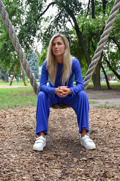 Attraktive blonde Frau, die auf einer hängenden Seilschaukel von einem Baum in einem blauen Anzug im Park sitzt