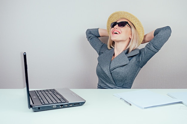 Attraktive blonde Frau (Geschäftsdame) im stylischen Business-Anzug, in Sonnenbrille und Strohhut träumt von Urlaub und Sonnenbaden im Büro mit Laptop auf dem Tisch. Reisekonzept und Urlaub