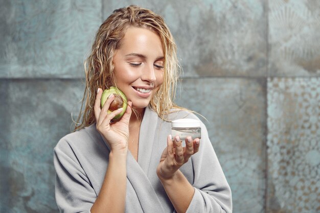 Attraktive blonde Frau hält nach dem Duschen Gesichtscreme und ein Stück Avocado im Badezimmer