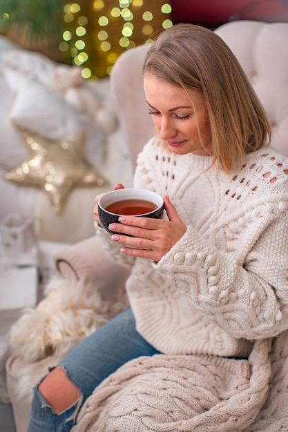 attraktive blonde Frau in einem gestrickten Pullover mit einer Tasse Tee sitzt auf dem Bett.