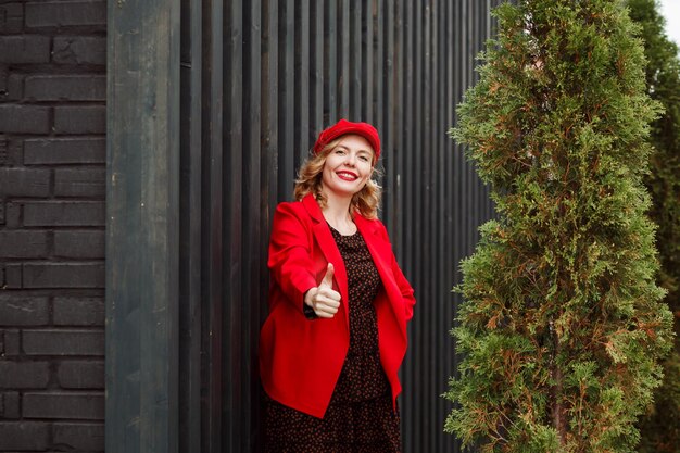 Attraktive blonde Frau in roter Jacke und Mütze, schwarzes Polka-Dot-Kleid zeigt Daumen hoch und posiert vor dunkler Holzwand, Street-Fashion-Konzept.