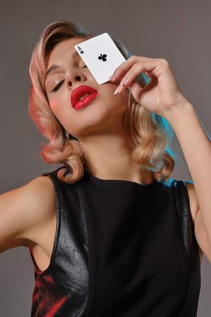 Attraktive blonde Frau mit hellem Make-up in schwarzem Lederkleid zeigt Kreuzass und posiert vor grauem Studiohintergrund Konzept des Glücksspiels Entertainment Poker Casino Closeup