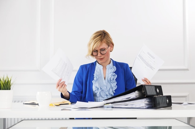 Attraktive blonde Geschäftsfrau trägt eine blaue Jacke, die mit Dokumenten auf dem Tisch mit einem beschäftigten Arbeitskonzept des weißen Büroraums im Blumentopf verwaltet wird