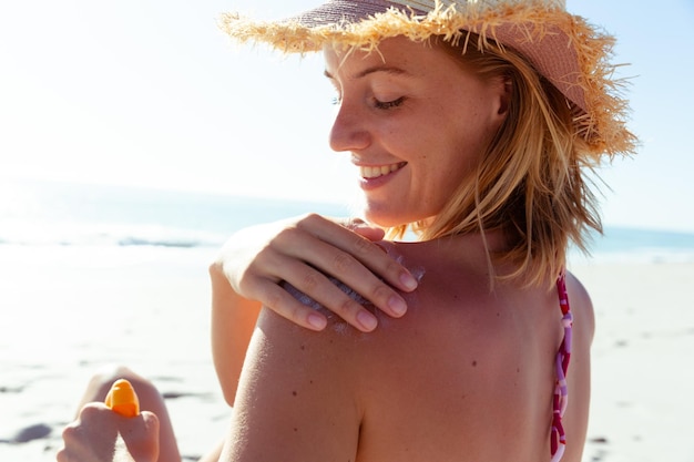 Attraktive blonde kaukasische Frau genießt die Zeit am Strand an einem sonnigen Tag und trägt Sonnencreme auf, mit blauem Himmel und Meer im Hintergrund. Sommerlicher tropischer Strandurlaub.