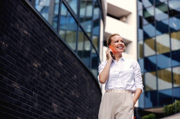 Attraktive blonde modische Geschäftsfrau, die am Telefon im Außenbereich des Geschäftszentrums spricht.