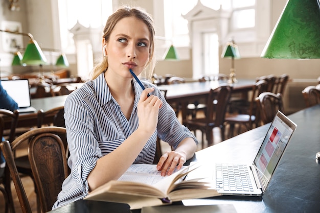 Attraktive blonde Studentin, die in der Bibliothek mit Laptop-Computer studiert