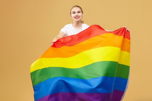 Attraktive Blondine posierte mit einer Regenbogen-LGBT-Flagge. Fotoshooting im Studio auf gelbem Hintergrund.