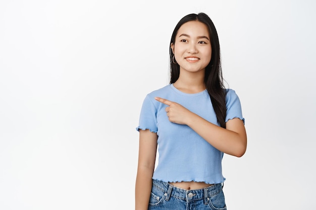 Attraktive brünette asiatische Frau, die auf die obere linke Ecke zeigt und das Promo-Angebot anstarrt, das im T-Shirt über weißem Hintergrund steht