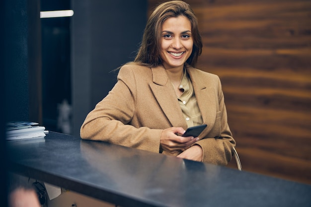 Attraktive brünette Frau in modischem Mantel, die mit ihrem Handy im Büro steht und lächelt