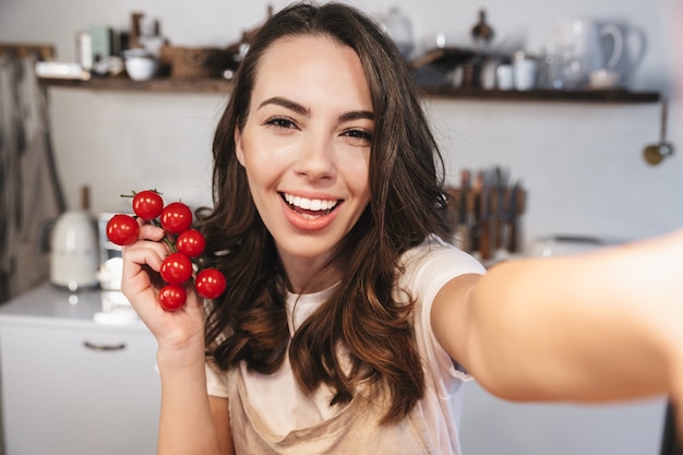 Attraktive brünette Frau mit Schürze, die ein Selfie macht, während sie zu Hause in der Küche sitzt und einen Haufen Tomaten hält