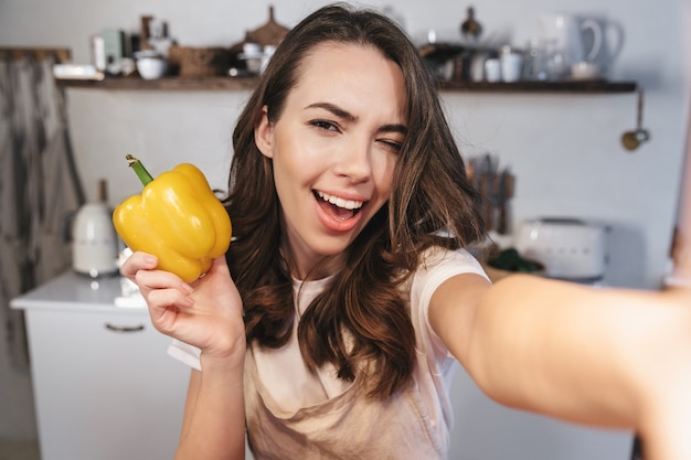 Attraktive brünette Frau mit Schürze, die ein Selfie macht, während sie zu Hause in der Küche sitzt und gelbe Paprika hält