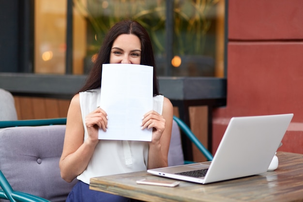 attraktive brünette Unternehmerin hält Papier in der Nähe Gesicht, lächelt positiv, verwendet Laptop-Computer für die Erstellung von Geschäftsberichten