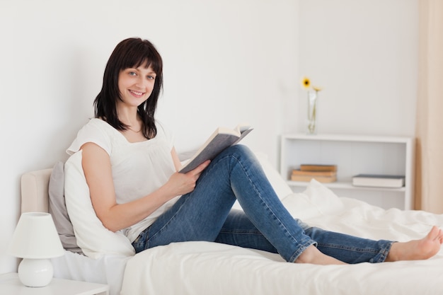 Attraktive Brunettefrau, die ein Buch beim Sitzen auf einem Bett liest