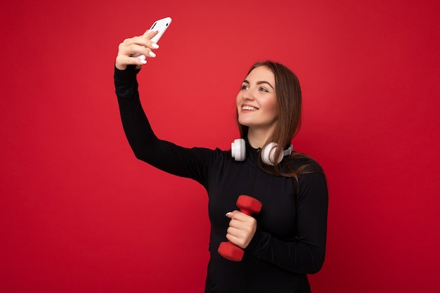 Attraktive charmante junge lächelnde glückliche Frau, die das Handy hält und benutzt, das selfie trägt, das stilvolle Kleidung einzeln über Wandhintergrund trägt.