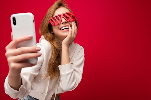 Attraktive charmante junge lächelnde glückliche Frau, die das Mobiltelefon hält und benutzt, das selfie trägt, das stilvolle Kleidung trägt, die über Wandhintergrund lokalisiert wird.