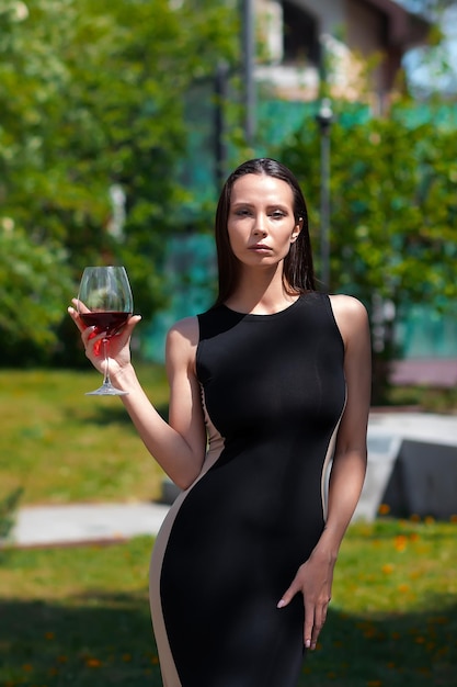 Attraktive Dame in schwarzem, elegantem Kleid, die den Hals berührt und an einem sonnigen Sommertag Rotwein genießt