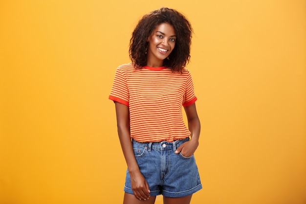 attraktive dunkelhäutige Frau mit lockigem Haar mit der Hand in der Tasche der Jeansshorts, die freudig lächelnd über orange Wand posiert