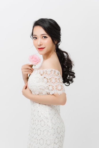 Attraktive elegante Asiatin, die Blume hält und weißes Kleid über weißem Hintergrund trägt