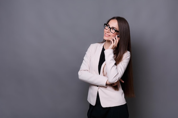Attraktive elegante Geschäftsfrau mit Brille, die auf ihrem Handy spricht und lächelt, während sie auf grauem Studiohintergrund mit Kopienraum steht. Geschäfts-, Technologie- und Kommunikationskonzept