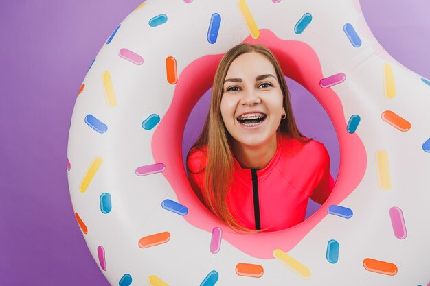 Attraktive emotionale Frau in stylischem rosafarbenem Badeanzug mit aufblasbarem Donut-Ring auf einfachem Hintergrund Strandmode