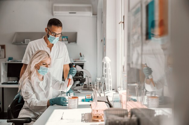 Attraktive engagierte blonde Laborassistentin mit Gesichtsmaske und Gummihandschuhen, die nach der Heilung des Koronavirus forscht.