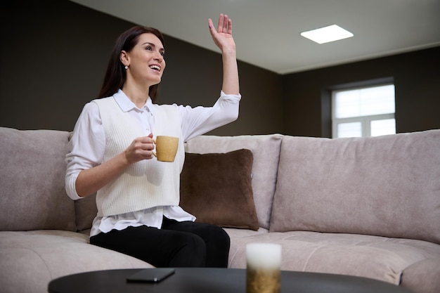 Attraktive, erfolgreiche, selbstbewusste Geschäftsfrau, die mit einer Tasse Kaffee auf einem Sofa im Ausstellungsraum eines Möbelhauses sitzt und während eines Geschäftstreffens lächelnd winkt und jemanden begrüßt