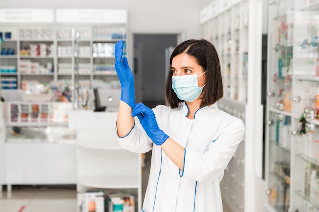 attraktive ernsthafte Apothekerin in der Apotheke mit Maske auf Gesicht, die blaue Handschuhe anzieht, um zu