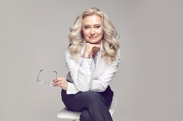 Attraktive erwachsene Dame mit blonden Haaren in einem stilvollen, lässigen Look, die auf einem Stuhl auf einem grauen, isolierten Hintergrund sitzt