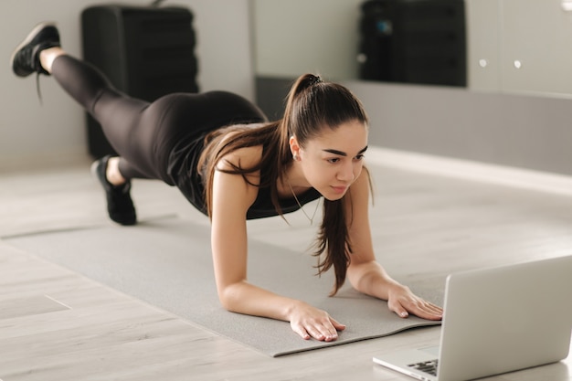 Attraktive fit Frau, die Yoga-Planke tut, während sie Online-Tutorials auf Laptop betrachtet. Ausbildung im Studium