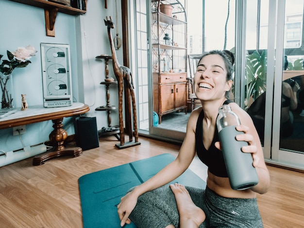 Attraktive Fitness junge Frau sitzt in Lotus-Pose Trinkwasser beim Yoga mit Online-Kurs zu Hause. Unterhaltung und Bildung im Internet. Gesundes Lifestyle-Konzept während der Freizeit.