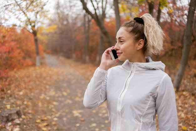 Attraktive Fitnessfrau in Sportkleidung, die im Herbstwald telefoniert