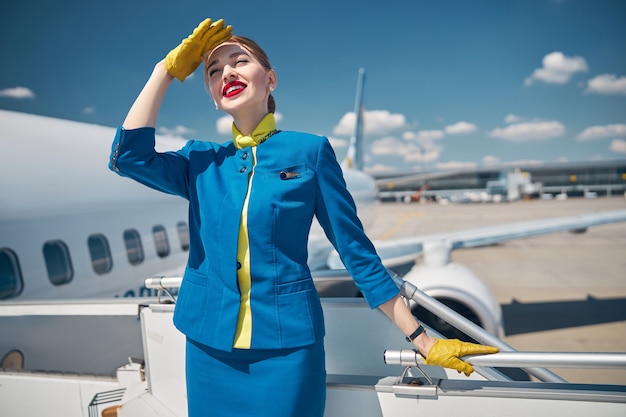 Attraktive flugbegleiterin in blauer uniform, die wegschaut und lächelt, während sie die augen vor der sonne mit der hand bedeckt