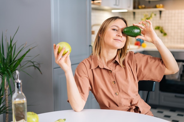 Attraktive Frau, die am Tisch sitzt und grüne, leckere Avocado vor Augen hält, in Küche mit modernem Interieur. Mädchen, das in der Hand Apfel hält. Konzept der gesunden Ernährung.