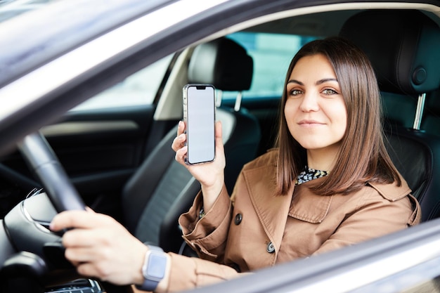 Attraktive Frau, die ein Auto fährt, zeigt einen leeren Telefonbildschirm und lächelt