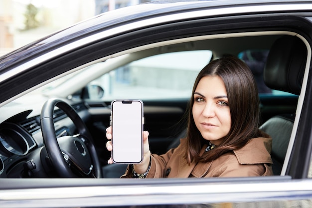 Attraktive Frau, die ein Auto fährt, zeigt einen leeren Telefonbildschirm und lächelt