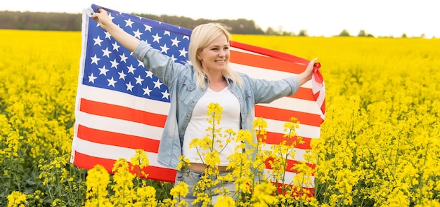 attraktive Frau, die eine amerikanische Flagge im Wind in einem Rapsfeld hält. Sommerlandschaft gegen den blauen Himmel. Horizontale Ausrichtung.