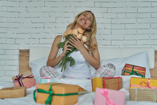 Attraktive Frau, die glücklich aussieht, während sie mit Geschenkboxen um sie herum im Bett sitzt